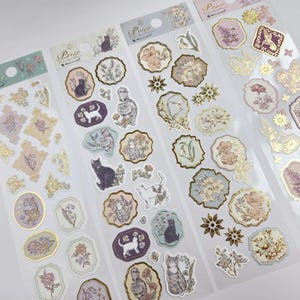 Op de afbeelding: Vellen decoratieve stickers met bloemen- en kattenillustraties in pastelkleuren met goudfolie accenten. De stickers bevatten verschillende vormen en maten, met het woord "Prior" bovenaan gedrukt.