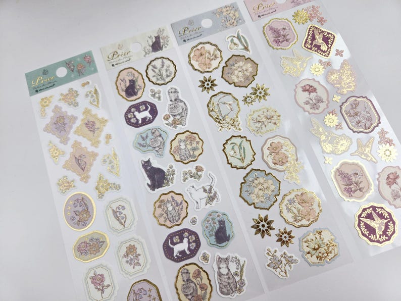 Op de afbeelding: Vellen decoratieve stickers met bloemen- en kattenillustraties in pastelkleuren met goudfolie accenten. De stickers bevatten verschillende vormen en maten, met het woord "Prior" bovenaan gedrukt.