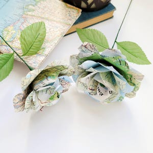 May include: Two paper roses made from vintage maps. The roses are light blue and green with a white background. The roses are on a white surface.