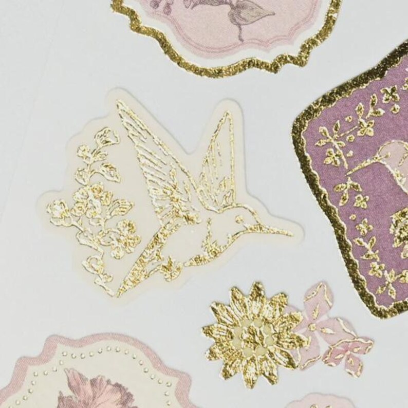 Op de afbeelding: Close-up van decoratieve stickers met gouden folie kolibries en bloemen ontwerpen. De stickers hebben een heldere, transparante achtergrond. Andere stickers hebben een roze en gouden kleurenschema, met een kolibrie en bloementhema.