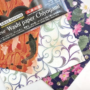 May include: A pack of Japanese Washi paper Chiyogami, with a black background and white text. The text reads "Japanese Washi paper Chiyogami" and "Career advancement". The pack contains three sheets of paper with different designs: one with red and gold koi fish, one with white and gold floral designs, and one with blue and green lotus flowers.