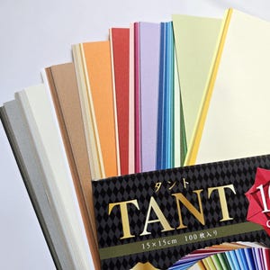 May include: A selection of colourful paper sheets in shades of grey, beige, orange, red, purple, blue, green, and yellow. The paper is 15 x 15 cm. The package features the word "TANT" in gold lettering on a black diamond pattern.