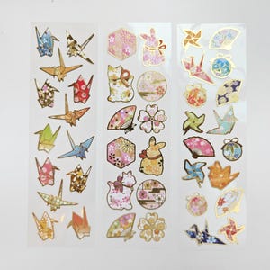 K&ouml;nnte beinhalten: Drei B&ouml;gen dekorativer Aufkleber mit Origami-Kranichen, F&auml;chern, Kaninchen und floralen Mustern. Die Aufkleber haben goldfarbene Akzente und sind in verschiedenen Farben, darunter Rosa, Blau und Gold.