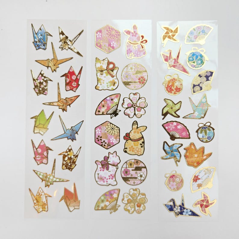 K&ouml;nnte beinhalten: Drei B&ouml;gen dekorativer Aufkleber mit Origami-Kranichen, F&auml;chern, Kaninchen und floralen Mustern. Die Aufkleber haben goldfarbene Akzente und sind in verschiedenen Farben, darunter Rosa, Blau und Gold.