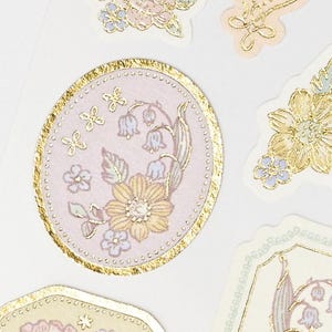 Op de afbeelding: Close-up van bloemenstickers met een gouden folierand. De stickers hebben pastelkleuren, waaronder roze, geel en blauw. De ontwerpen omvatten bloemen, vlinders en andere botanische elementen. De stickers zijn gerangschikt op een witte achtergrond.