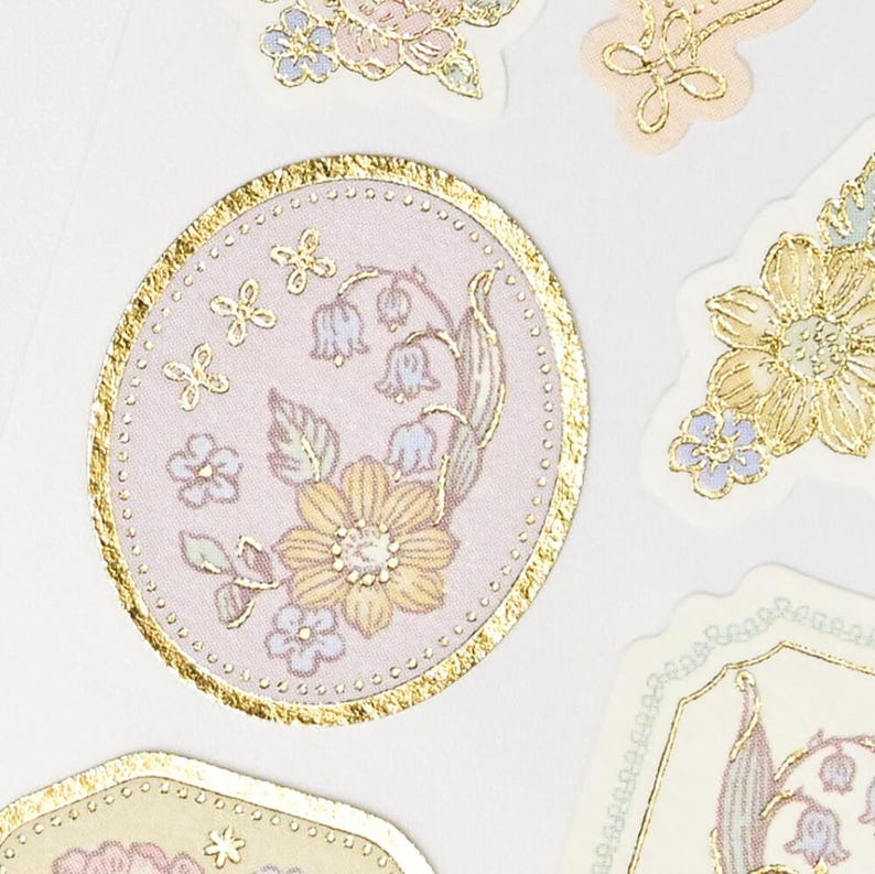 Op de afbeelding: Close-up van bloemenstickers met een gouden folierand. De stickers hebben pastelkleuren, waaronder roze, geel en blauw. De ontwerpen omvatten bloemen, vlinders en andere botanische elementen. De stickers zijn gerangschikt op een witte achtergrond.