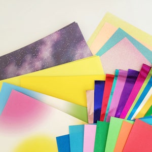 May include: A selection of colourful paper sheets, including iridescent, glitter, and patterned designs. The papers are arranged in a fan shape, with the top sheet being a dark purple with a galaxy pattern.
