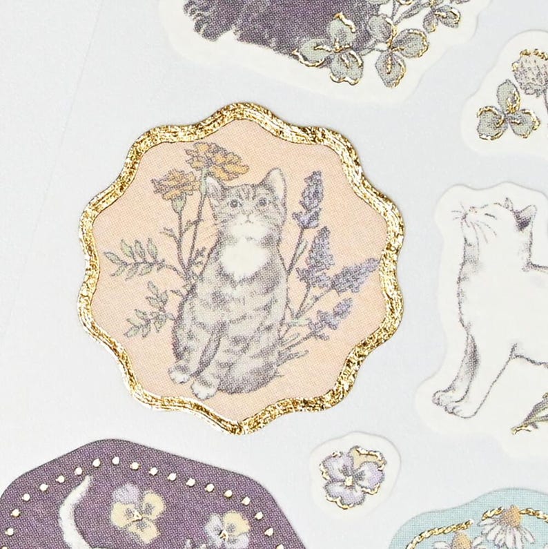 Op de afbeelding: Een verzameling decoratieve stickers met katten- en bloemenillustraties. De centrale sticker toont een grijze tabby-kat omringd door bloemen, omlijst met een gouden rand. Andere stickers bevatten een witte kat en diverse bloemmotieven.