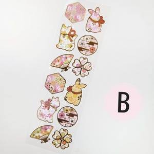 K&ouml;nnte beinhalten: Ein Streifen dekorativer Aufkleber mit japanisch inspirierten Designs. Die Aufkleber zeigen Katzen, Kaninchen, F&auml;cher, Blumen und geometrische Formen, die mit floralen Mustern in Rosa, Gold und Wei&szlig; verziert sind. Ein rosa Kreis mit dem Buchstaben "B" befindet sich in der unteren rechten Ecke.