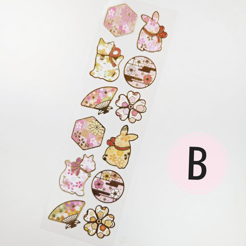 K&ouml;nnte beinhalten: Ein Streifen dekorativer Aufkleber mit japanisch inspirierten Designs. Die Aufkleber zeigen Katzen, Kaninchen, F&auml;cher, Blumen und geometrische Formen, die mit floralen Mustern in Rosa, Gold und Wei&szlig; verziert sind. Ein rosa Kreis mit dem Buchstaben "B" befindet sich in der unteren rechten Ecke.
