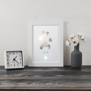 Puede incluir: Una imagen enmarcada en blanco con una huella de pata plateada y el nombre "Cash" escrito debajo. La imagen está sobre una mesa de madera con un reloj blanco y un jarrón de flores blancas.