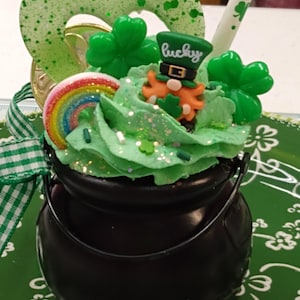 May include: A black pot-shaped dessert with green frosting, shamrocks, a rainbow, and a leprechaun figurine. A heart-shaped decoration and a straw with shamrock designs are also present. The dessert is decorated with glitter and sprinkles, and the word "lucky" is visible.