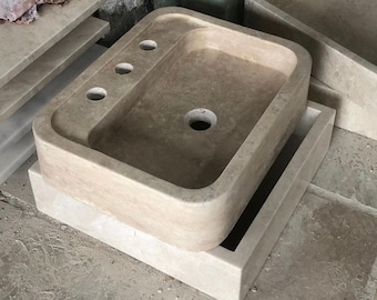 May include: A beige stone bathroom sink with three holes for faucets and a drain hole. The sink is sitting on a beige stone base.