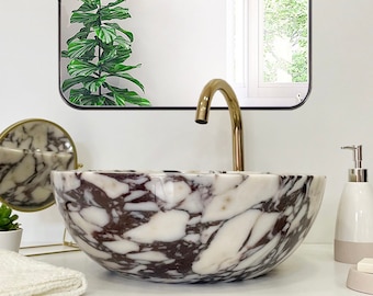 May include: A white marble vessel sink with brown and gray veining. The sink is on a white countertop with a gold trim. A gold faucet is mounted above the sink. A small potted plant, a white towel, and a white soap dispenser are also on the countertop. A mirror with the text "THE MERCI DESIGN" is mounted above the sink.