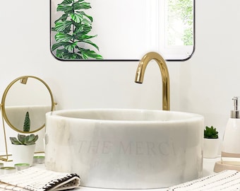 May include: A bathroom vanity with a white marble sink, gold faucet, and mirror. The mirror reflects a green plant. The sink has the text "THE MERCI DESIGN". Two black and white patterned towels are on the counter.