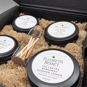 May include: A gift set featuring scented candles and matches. The candles are in black tins with white labels, and the scents are named "Netherfield Ball", "Mr. Darcy", and "Elizabeth Bennet". The matches are in a glass vial.