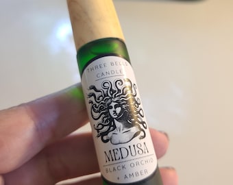 Pode incluir: Um frasco roll-on de vidro verde com tampa de madeira. O rótulo diz "THREE BELLS CANDLE" e apresenta uma ilustração em preto e branco de Medusa. O texto abaixo diz "MEDUSA BLACK ORCHID + AMBER."