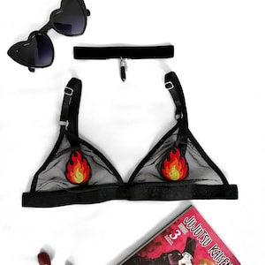 Op de afbeelding: Een zwarte lingerie set met een doorschijnende driehoekige bh met vlammenpatches, een zwarte fluwelen choker met een hanger en hartvormige zonnebrillen. Een exemplaar van de manga "Jujutsu Kaisen" is ook aanwezig.