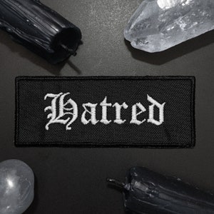 Puede incluir: Parche de tela negro con texto bordado en blanco que dice "Hatred" en una fuente gótica.