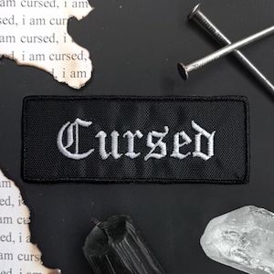 Może przedstawiać: Czarny materiałowy naszywka z białym haftem, na którym widnieje napis "Cursed" w staroangielskim stylu.