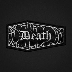 May include: Black patch with white embroidered text that reads "Death" in a gothic font. The patch is rectangular with a black background and a white spiderweb design around the edges.