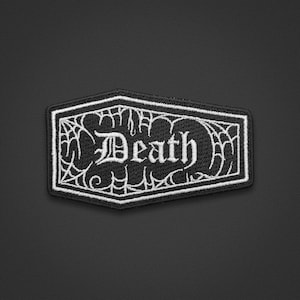 以下が含まれることがあります： 棺桶の形をした黒と白の刺繍ワッペン。ワッペンには、ゴシック体のフォントで書かれたDeathという文字を囲む白いクモの巣模様が施されており、全体が黒い布地の背景に白い縁取りで囲まれています。