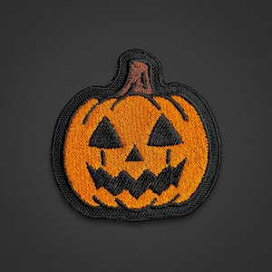 May include: An embroidered patch featuring a jack-o'-lantern pumpkin design. The pumpkin is bright orange with a brown stem and black thread detailing for the triangular eyes, small triangular nose, and a jagged, toothy grin, all outlined in black.