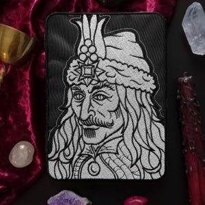 Può includere: Toppa rettangolare nera con un ritratto dettagliato in scala di grigi di una figura storica con baffi e copricapo ornato. La toppa ha uno sfondo nero e un design ricamato bianco, adatta per giacche o borse.