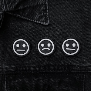 May include: Three black and white embroidered patches with different facial expressions: two neutral and one sad. The patches are circular and feature a white outline and details. The patches are on a black denim jacket.