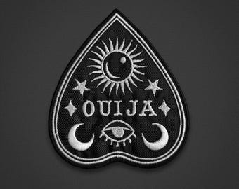 May include: A heart-shaped black embroidered patch featuring white thread details. The design includes a central sun, the word OUIJA, two stars, two crescent moons, and an eye symbol. The patch has a white border and a textured fabric finish.