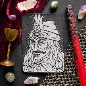 Puede incluir: Un parche bordado rectangular que presenta un retrato detallado en hilo blanco de una figura histórica con cabello largo, bigote y un sombrero alto y ornamentado sobre un fondo negro. El diseño incluye líneas intrincadas y un colgante.