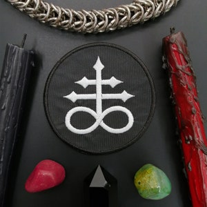 Puede incluir: Un parche negro con un símbolo blanco de una cruz con un símbolo de infinito debajo. El parche está rodeado por una cadena plateada, dos velas negras, una vela roja, una piedra roja y una piedra verde.