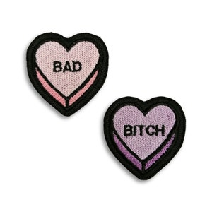 Pode incluir: Dois patches bordados em forma de coração com contornos pretos. O patch superior é rosa e diz "BAD" em preto. O patch inferior é roxo e diz "BITCH" em preto.
