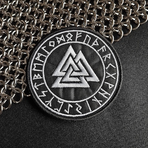 May include: A circular black embroidered patch featuring a white Valknut symbol at the centre, surrounded by a border of white runic characters. The patch is stitched with high-contrast thread and has a clean, defined edge, showcasing intricate needlework.