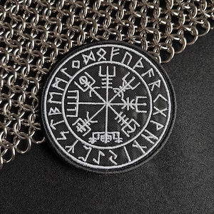 May include: A circular black embroidered patch featuring a white Vegvisir symbol at the center, surrounded by a border of runic characters. The patch is positioned against a background of interlocking silver-colored metal chainmail links.