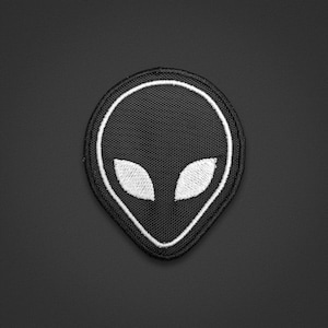 May include: A circular embroidered patch featuring a black background with a white stitched outline of an alien head shape. The design includes two large, almond-shaped white eyes centred within the black field, all finished with a black border.