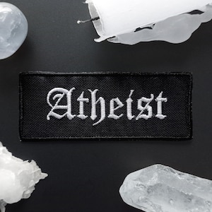 May include: Black rectangular patch with the word "Atheist" embroidered in white gothic lettering. The patch is surrounded by a black background with a white candle and crystals.