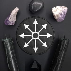 May include: A black patch with a white eight-pointed star with arrows pointing outwards. The patch is surrounded by black candles, amethyst crystals, and black stones.