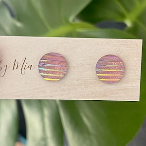 Könnte beinhalten: Ein Paar Ohrstecker in irisierendem Rosa und Gold mit Streifen. Die Ohrstecker liegen auf einer hellbraunen Holzoberfläche mit dem Schriftzug "by Mia".