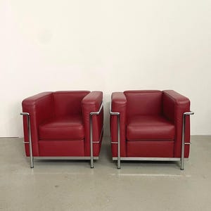 Könnte beinhalten: Zwei rote Ledersessel mit verchromten Gestellen. Die Stühle haben ein kastenförmiges Design mit quadratischen Kissen und Armlehnen. Die verchromten Gestelle bilden einen modernen Kontrast zum roten Leder. Die Stühle stehen nebeneinander.