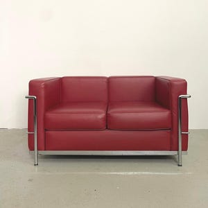 Könnte beinhalten: Ein rotes Ledersofa für zwei Personen mit minimalistischem Design. Das Sofa hat eine kastenförmige Form mit Armlehnen und einem Gestell aus verchromtem Stahlrohr. Die Kissen sind tiefrot, und das Sofa steht vor einer schlichten weißen Wand.