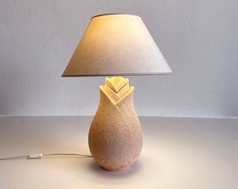 May include: A beige ceramic table lamp with a white fabric lampshade. The lamp base is shaped like a flower bud with a textured surface.