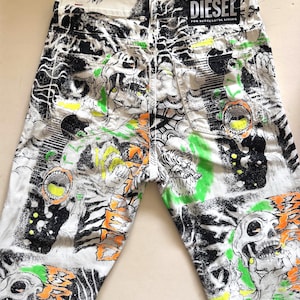 Puede incluir: Vaqueros de mezclilla blancos con un estampado gráfico llamativo en toda la superficie que presenta calaveras, tigres y diseños abstractos en negro, verde y naranja. Los vaqueros tienen una etiqueta Diesel en la cintura.