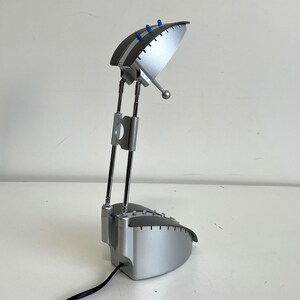 Può includere: Una lampada da scrivania argentata con paralume curvo e bracci regolabili. La lampada ha un design moderno con accenti blu sul paralume e finitura cromata sui bracci. È collegata a un cavo di alimentazione nero.