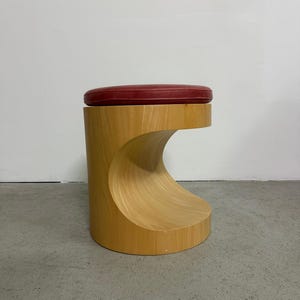 Puede incluir: Un taburete de madera moderno con un diseño curvo. Cuenta con un asiento circular de cuero rojo. La base es de madera clara, con un recorte en forma de ola. Este mueble es adecuado para una sala de estar o un dormitorio.
