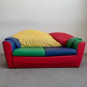 Op de afbeelding: Een kleurrijke tweezitsbank met een levendig ontwerp. De bank heeft een rode basis, met groene en blauwe zitkussens. De rugkussens zijn geel en rood, en de armleuningen zijn blauw en groen. Een speels en modern meubelstuk.