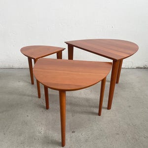 Puede incluir: Juego de tres mesas nido de estilo moderno de mediados de siglo. Cada mesa tiene una tapa semicircular y patas cónicas. Las mesas son de madera con un acabado marrón rojizo cálido. La mesa más grande mide aproximadamente 76 cm de ancho.