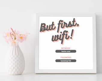 May include: A framed white print with the text "But first, wifi!" in a playful font. Below, the network name "hecho cool" and password "bestshop123" are displayed. A white vase with pink flowers sits to the left.