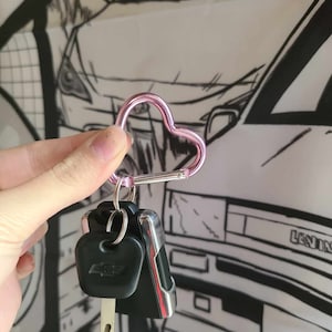 Puede incluir: Un llavero de mosquetón en forma de corazón rosa con una llave de coche negra adjunta. El llavero se sostiene en una mano sobre un fondo blanco con un dibujo en blanco y negro de un coche.