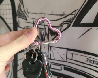 May include: A pink heart-shaped carabiner keychain with a black car key attached. The keychain is held in a hand against a white background with a black and white drawing of a car.